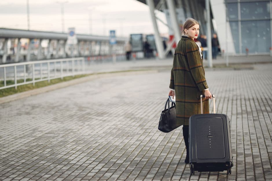Full length of stylish female traveler in warm coat walking with suitcase and bag looking at camera after arrival against airport terminal