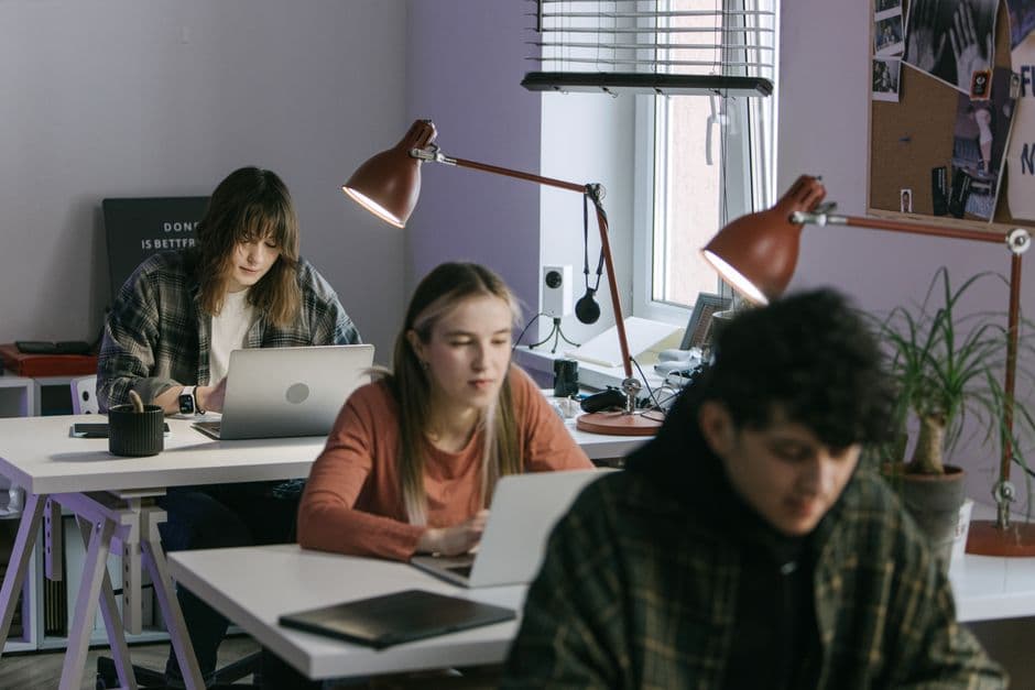 Three young professionals working at desks in a modern office setting with laptops and desk lamps.
