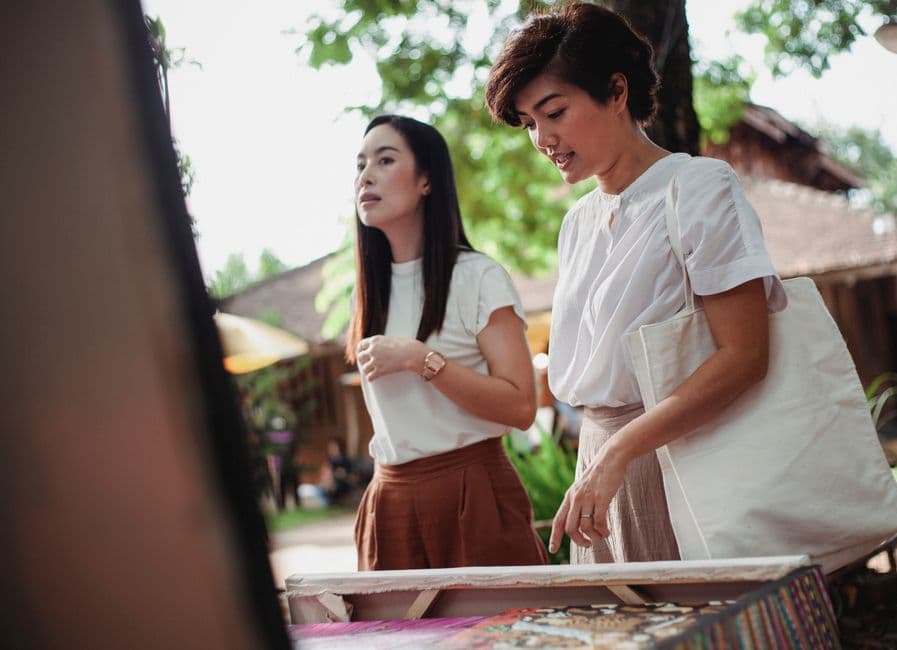 Positive young Asian females in stylish summer outfits standing near stall with souvenirs and choosing gifts while spending sunny day in street market