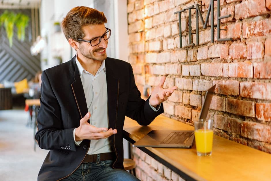 Smiling man in black jacket gesturing during a video call in a modern cafe.