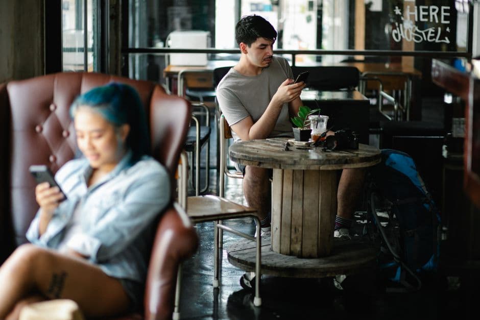 Focused male text messaging on cellphone while sitting at table with refreshing beverage in light cafe with blurred Asian woman