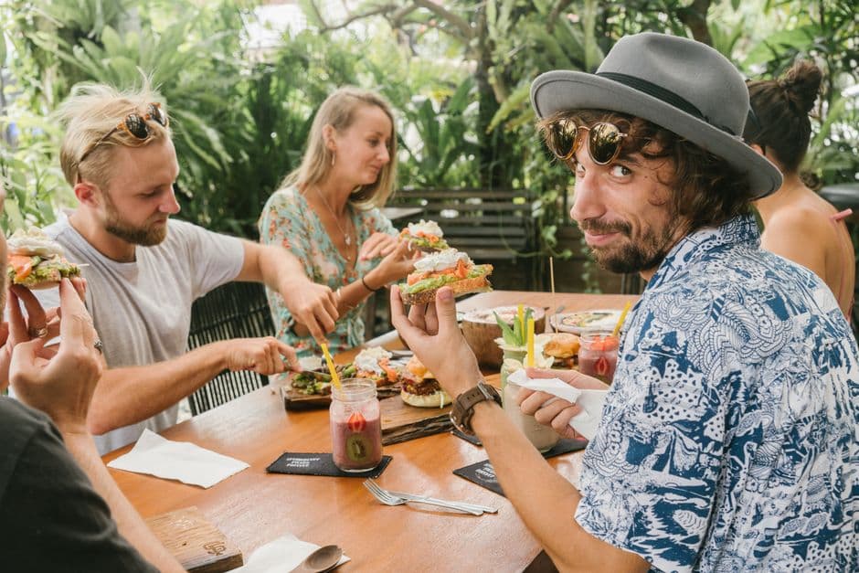 Friends gather around a table enjoying brunch outdoors with sandwiches and drinks.