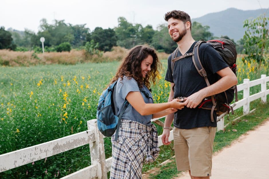 Side view of cheerful young female hiker in casual clothes fastening backpack of smiling boyfriend while standing together near wooden fence and lush green meadow during hiking in mountainous countrys