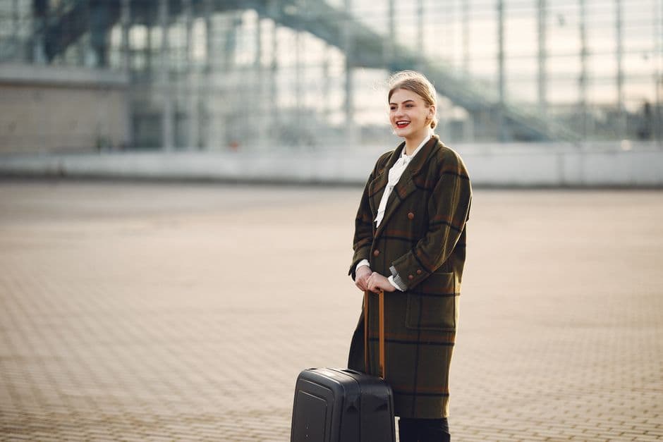 Side view of cheerful stylish blond woman in casual wear with big suitcase standing against airport and waiting for flight