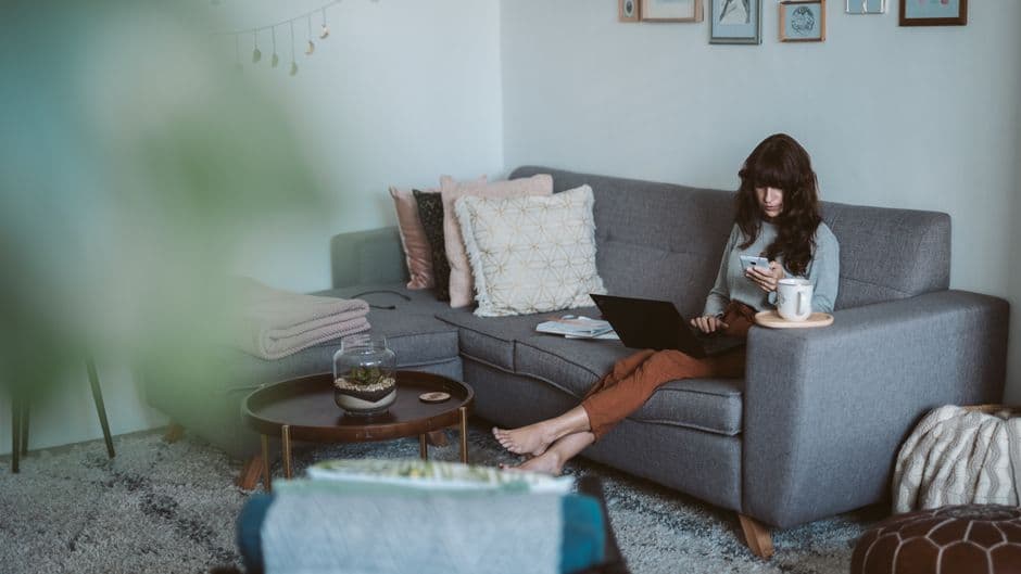 Woman working from home on a sofa with a laptop and phone, in a cozy living room setting.