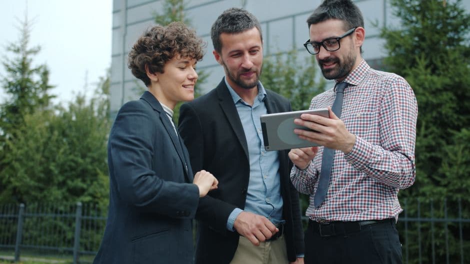 Three professionals engaged in a discussion while viewing a tablet outdoors.