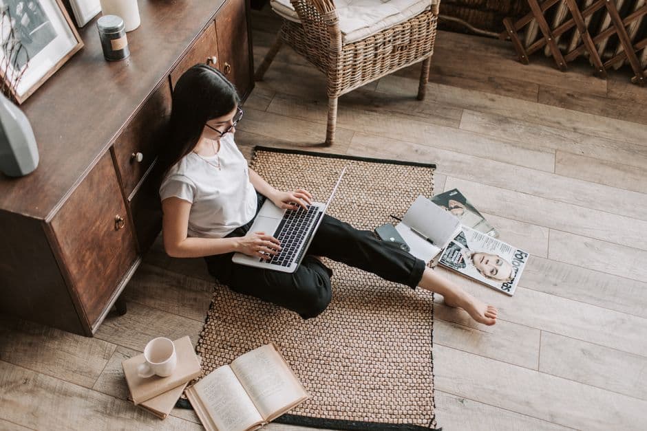 From above of female freelancer using netbook while sitting on floor carpet near chest of drawers and wicker chair among magazines and books during remote work on project at home